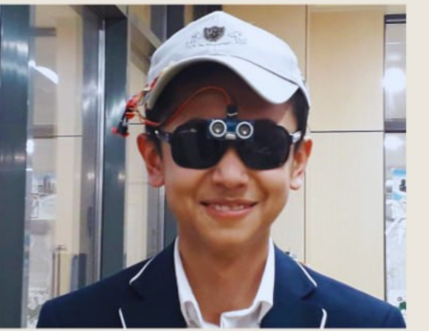 Assistive device: person wearing sunglasses with sensor module, baseball cap with electronics, dark blue blazer with white piping